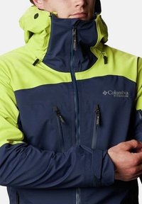 Giacca blu navy e giallo brillante con colletto alto, tasche con zip sul petto e cappuccio regolabile per l'uso all'aperto. Logo Columbia Titanium visibile.