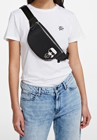 Czarna nylonowa torba na ramię z zamkiem błyskawicznym, z białym kreskówkowym nadrukiem i logo. Noszona na skos przez ciało, w połączeniu z białym T-shirtem i dżinsami.