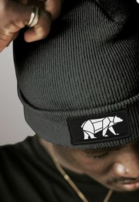 Person wearing a dark ribbed knit beanie with a geometric white bear patch, adjusting it with a hand showing a silver ring.