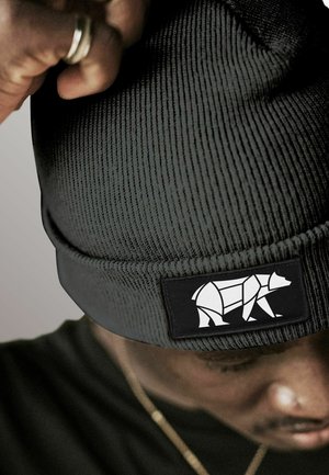 Person wearing a dark ribbed knit beanie with a geometric white bear patch, adjusting it with a hand showing a silver ring.