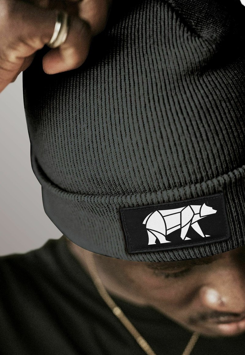 Person wearing a dark ribbed knit beanie with a geometric white bear patch, adjusting it with a hand showing a silver ring.