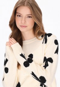 Krämfärgad stickad tröja med stora svarta prickar och rosetter, långa ärmar och en rund halsringning med subtila glittrande detaljer.