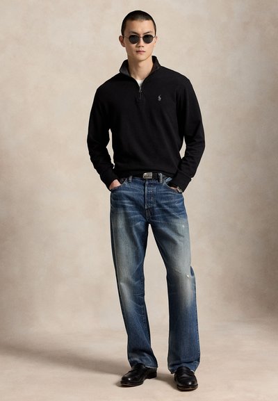 Hombre con suéter negro con cremallera hasta el cuarto, jeans azules, zapatos negros y gafas de sol redondas, de pie con las manos en los bolsillos sobre fondo beige.