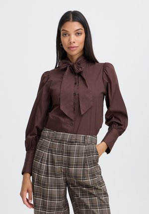Femme aux longs cheveux foncés, portant un chemisier marron avec un grand nœud au cou et un pantalon à carreaux taille haute, debout devant un fond uni.