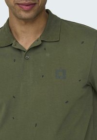 Olivgrünes Poloshirt aus weichem Baumwollstoff, mit einem Drei-Knopf-Verschluss, kleinen Blattmuster und einem gebrandeten Grafik in einer dunkleren Farbtönung.