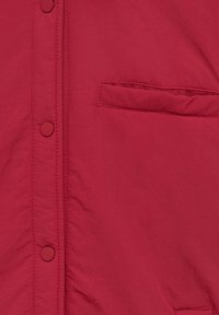Veste légère rouge avec une patte de boutonnage à boutons-pression sur le devant et une poche poitrine unique. Tissu lisse, légèrement texturé.