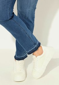 Bílé plátěné tenisky s texturovaným povrchem, nosené s modrými džíny s roztrhanými nohavicemi a se štíhlým, rovně střiženým střihem.