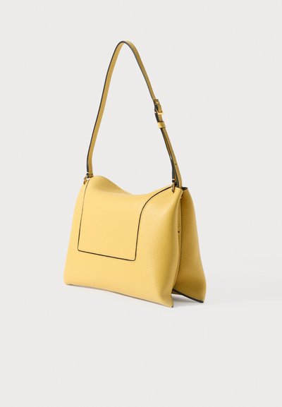 Dzeltena teksturēta ādas plecu soma ar regulējamu siksnu un melnu apmali uz vienkrāsaina balta fona.