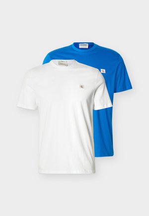 Două tricouri simple cu guler rotund, unul alb și unul albastru, fiecare cu o mică emblemă cu sigla Calvin Klein pe piept.
