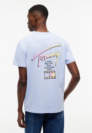 Mann trägt hellblaues T-Shirt mit pastellfarbenem "Tommy"-Logo und Städtenamen, kombiniert mit blauen Jeans, von hinten vor weißem Hintergrund gezeigt.