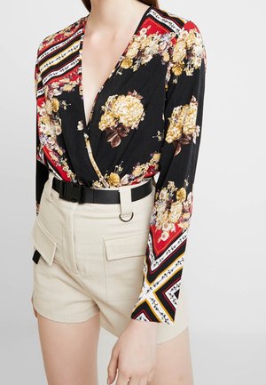 Blouse portefeuille à manches longues avec motif floral rouge, noir et jaune, associée à un short beige taille haute et une ceinture noire.