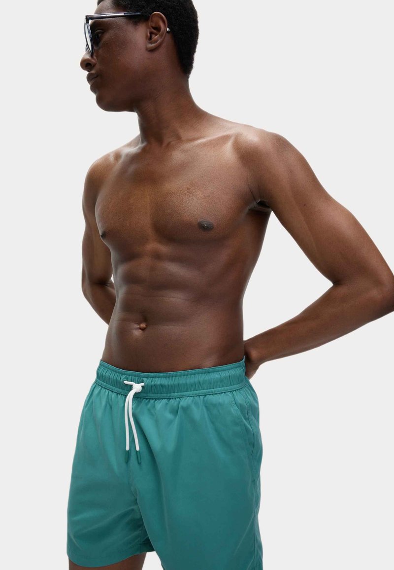Turkusowe szorty kąpielowe z elastycznym pasem i białym sznurkiem, wykonane z lekkiego materiału, charakteryzujące się gładką teksturą.