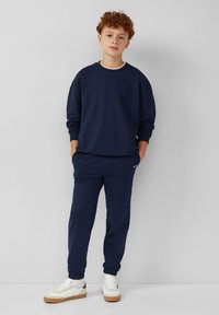 Navy sweatshirt en bijpassende joggers van zachte stof, met ribbelmanchetten en een klein logodetail. Gecombineerd met witte sneakers.