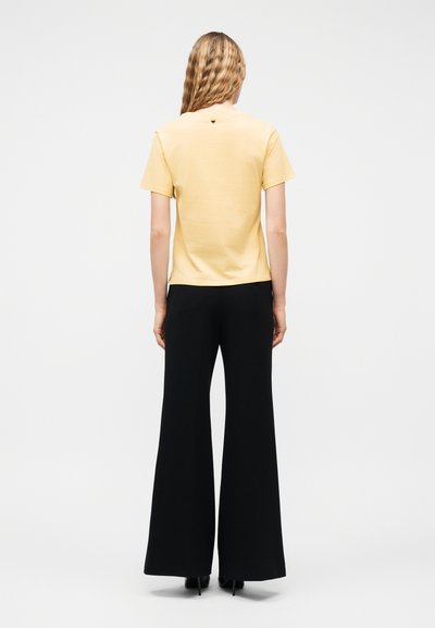 Femme aux cheveux blonds ondulés portant un chemisier jaune pâle à manches courtes et un pantalon noir à jambes larges, debout de dos sur un fond uni.