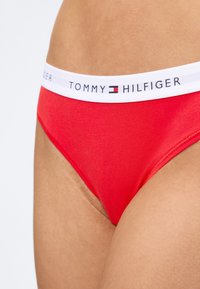 Gros plan sur une personne portant un slip rouge Tommy Hilfiger avec une ceinture blanche arborant le nom et le logo de la marque.