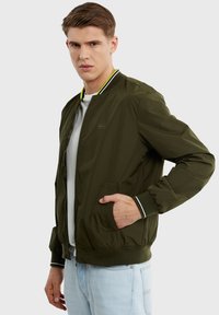 Alyvuogių žalia bomber tipo striukė, nailono medžiaga, kantai ir apatinė dalis su baltais dryžiais, šoninės kišenės ir minimalistinis logotipo akcentas.