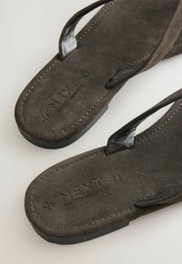 Rudos zomšos sandalai su juodais diržais, su tekstūruotais padais ir įspaudu „NEXT“ ant pado, ilsinčiais ant šviesaus fono.