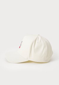 Hvit baseballcap med buet skjerm, sydd detaljer og en brodert design på fronten. Laget av bomullsstoff.