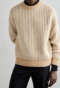 Mann in einem cremefarbenen, strukturierten Strickpullover über einem schwarzen Hemd, kombiniert mit schwarzen Hosen, steht vor einem schlichten hellen Hintergrund.