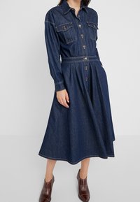 Jeanshemdkleid in Dunkelblau, mit einem kragenförmigen Ausschnitt, durchgehender Knopfleiste, zwei Brusttaschen und einem taillierten Bund, der in einen Midi-Saum übergeht.