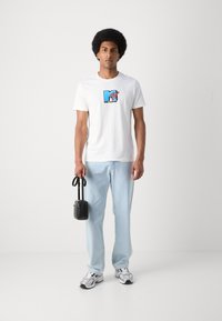Camiseta de algodón blanca con un logo de MTV en azul y rojo, combinada con jeans de tonalidad azul clara y zapatillas plateadas, sosteniendo un bolso cruzado negro.
