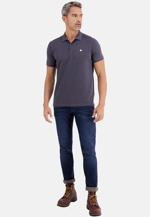 Uomo in piedi che indossa una polo grigio scuro, jeans blu scuro risvoltati e stivali marroni con lacci, che guarda verso sinistra.