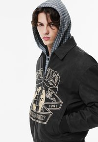 Musta nahkatakki, jossa on etuvetoketju, kermanvärinen graafinen painatus, sivutaskut ja harmaa ruudullinen hupullinen vuori.