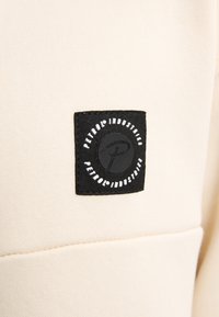 Tessuto beige con una texture liscia, caratterizzato da un'etichetta nera quadrata. L'etichetta presenta testo circolare bianco e un logo stilizzato al centro.