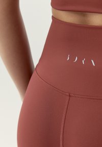 Bordové legíny s vysokým pásom vyrobené z hladkej, elastickej tkaniny s jemným leskom. Obsahujú viditeľný detail bieleho loga a bezšvový dizajn.