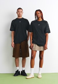 Two individuals stand side by side, wearing black athletic t-shirts with logo, brown cargo shorts, and light-colored skirts. White socks and sneakers are visible.