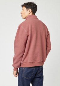 Fleece pullover in gedempt roze met een hoge kraag, gestructureerde buitenkant en ribgebreide zoom. Gecombineerd met donkerblauwe denim jeans.