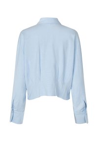 Světle modrá zkrácená košile s límečkem a dlouhými rukávy. Má jemnou texturu a knoflíkové manžety, s bočními záhyby pro uvolněný střih.