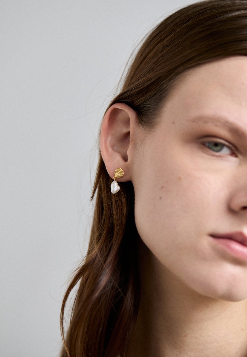 Cercei aurii cu un disc texturat și o perlă albă de apă dulce care atârnă. Design minimalist cu un finisaj natural.