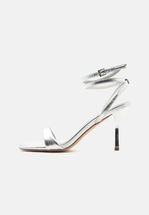 Sølv høye hæler sandal med ankelbånd og tynn stilettohæl, vist fra siden på hvit bakgrunn.