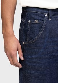 Jeans din denim albastru închis cu o textură subtilă, cu detalii metalice argintii și un logo pe buzunar. Vedere laterală care arată o mână sprijinită pe țesătură.