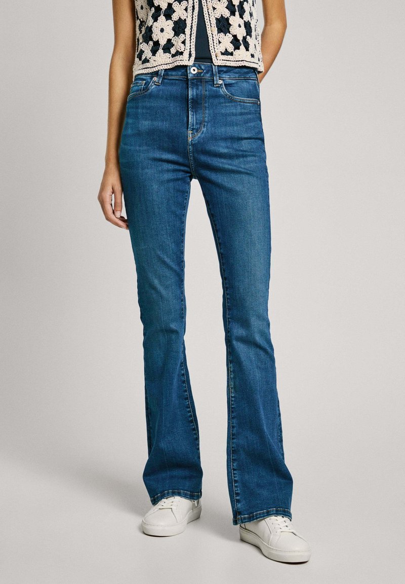 Blå utsvängda jeans i denim, med en klassisk fem-fickorsdesign, medelhög midja och kontrasterande sömmar. Vita sneakers bärs.