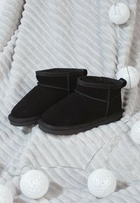 Botines de ante negros con un suave forro interior, que presentan un detalle de costura y una suela de goma plana, colocados sobre una manta texturizada.