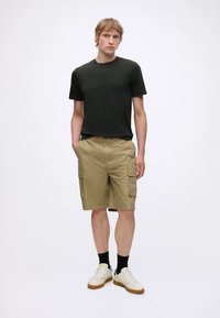 Jeune homme debout portant un t-shirt noir, un short cargo kaki, des chaussettes noires et des baskets blanches sur fond blanc uni.