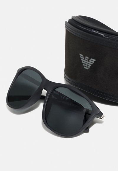 Occhiali da sole neri oversize con lenti grigie e finitura opaca, accompagnati da una custodia nera con logo argento.