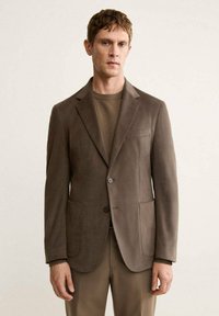 Brown, textured blazer with two front pockets, single button closure, tailored fit, worn over a brown sweater. Simple design, no patterns.