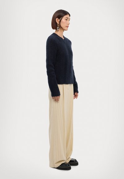 Suéter de punto azul marino con un patrón de canalé vertical, combinado con pantalones crema a rayas y zapatos negros de suela gruesa.
