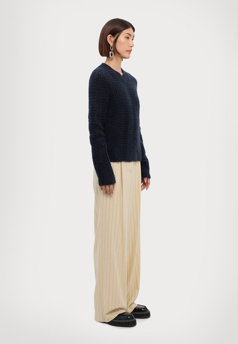Navy knitted sweater with a vertical rib pattern, paired with cream pinstriped trousers and black chunky-soled shoes.