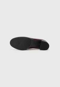 Chaussure bordeaux avec une tige en cuir lisse, un bout arrondi et une semelle en caoutchouc noire texturée. Design minimaliste sans accents visibles.