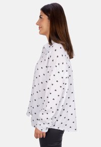 White long-sleeve blouse with black heart patterns, relaxed fit, textured fabric, and buttoned cuffs. Showcased from the side.