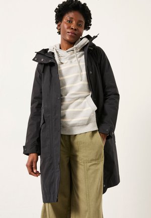 Black woman wearing black hooded coat over gray striped hoodie and olive wide-leg pants, standing against a plain light background.