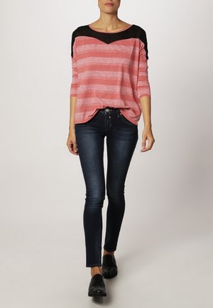 Kvinde går iført en pink og sort stribet langærmet top, mørke skinny jeans og sorte nittede sko mod en ensfarvet baggrund.