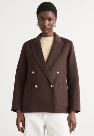 Brown double-breasted wool blazer with gold buttons, notched collar, and two front pockets, paired with a light beige top.