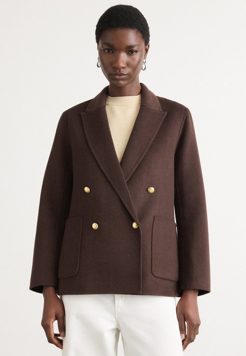 Bruin dubbelgebreid wollen blazer met gouden knopen, geknopt kraag en twee voorzakken, gecombineerd met een lichtbeige top.