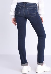 Jeans skinny in denim blu scuro, con due tasche posteriori con cuciture a contrasto, orli arrotolati e abbinati a sneakers bianche.