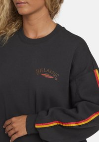 Sudadera negra con un logo en el frontal con acentos rojos y amarillos, y detalles a rayas en las mangas en rojo y amarillo. Tejido texturizado.